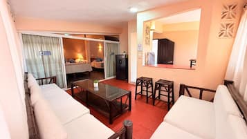 Un salon avec une table basse en verre, des canapés blancs, une petite table à manger avec des chaises et une porte coulissante donnant sur une chambre.
