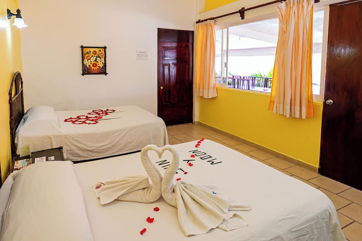 A hotel room with two beds, each with a heart-shaped towel arrangement and red rose petals. There is a painting on the wall and a window with curtains.