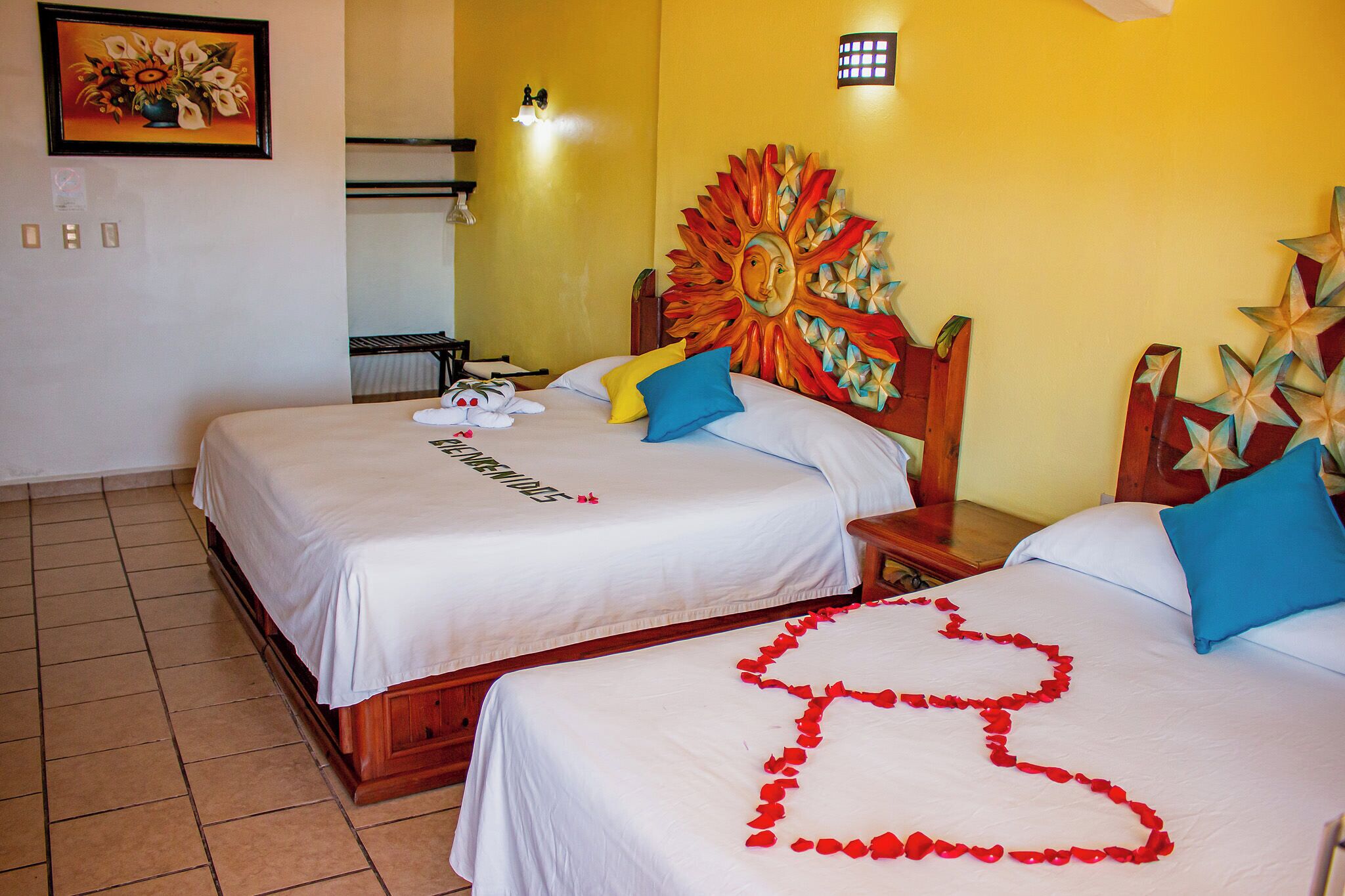 A hotel room with two beds, a decorative sun on the headboard, and a heart-shaped rose petal design on the bed.