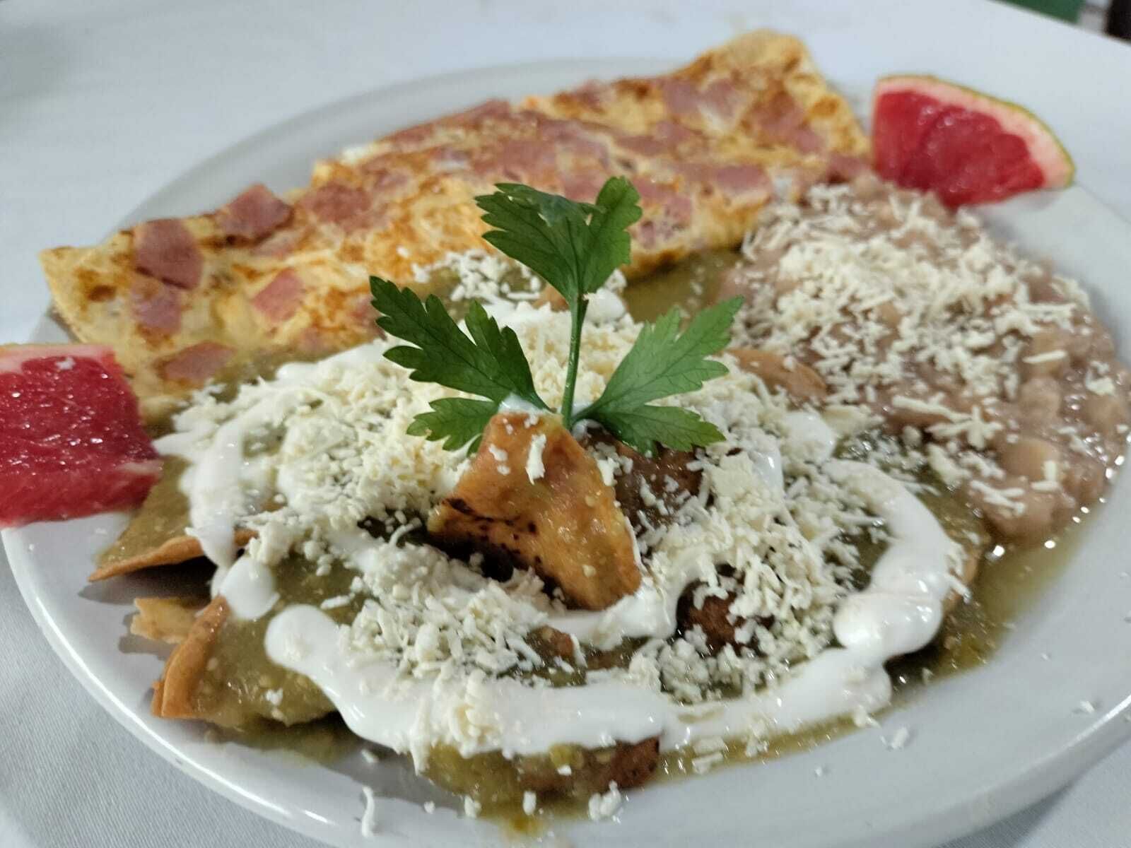 A plate of food with a garnish of parsley, including a dish with a creamy sauce and cheese, a slice of bread, and a piece of fruit.