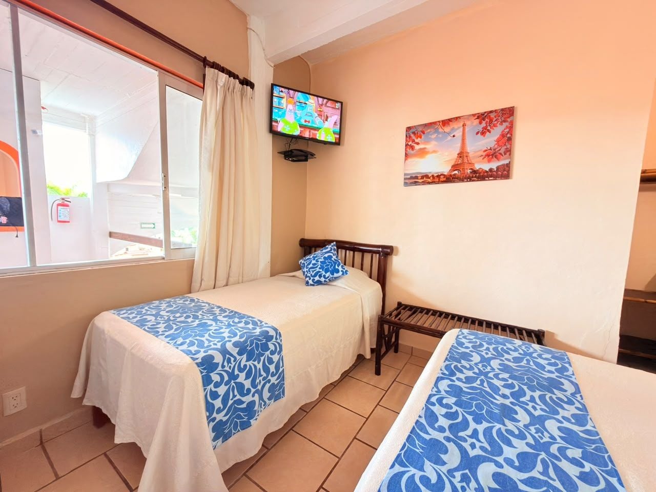 A hotel room with two beds, a television, a painting of the Eiffel Tower, and a window with curtains.