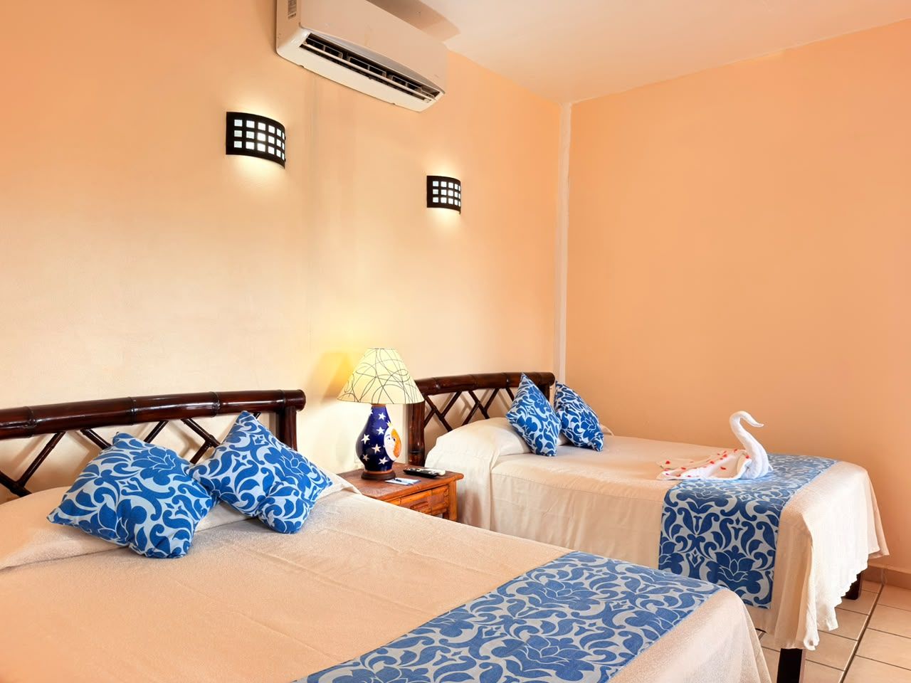 Habitación de hotel con dos camas, cada una con almohadones azules estampados a juego, una mesita de noche con lámpara y un cisne decorativo sobre la cama.
