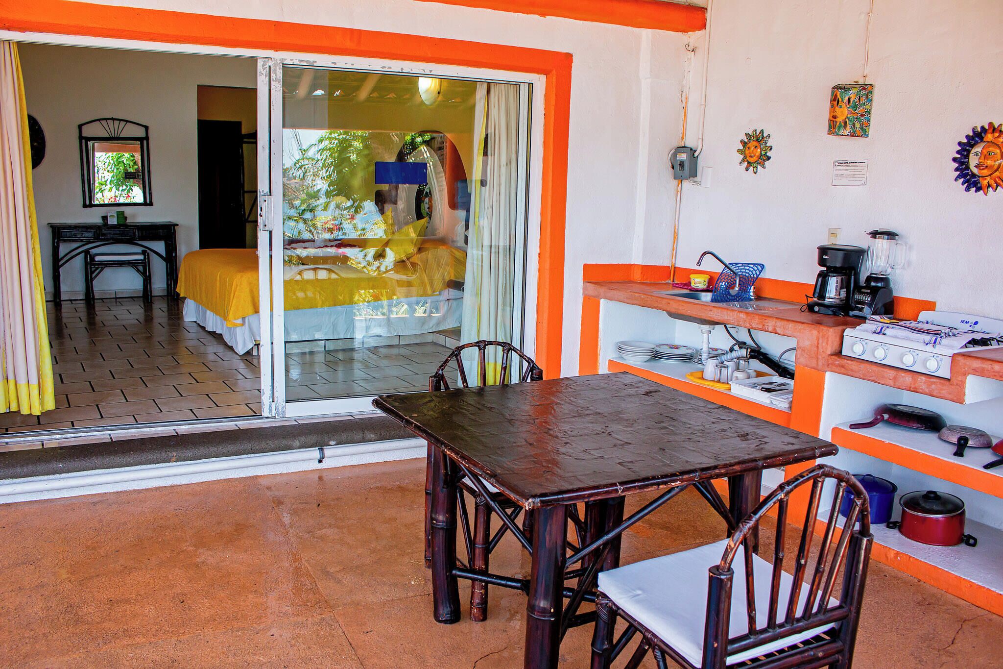 A room with a sliding glass door leading to a balcony, a wooden table with chairs, a kitchen area with a stove and various kitchen items, and a bed with a yellow blanket.
