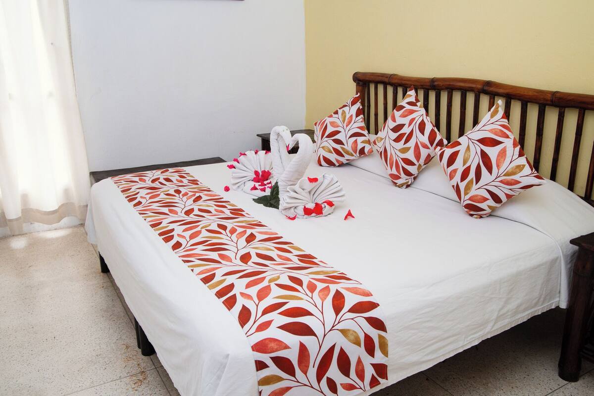 Una cama bien tendida con una colcha blanca y almohadas estampadas con hojas rojas, un cabecero de madera y una mesita de noche.