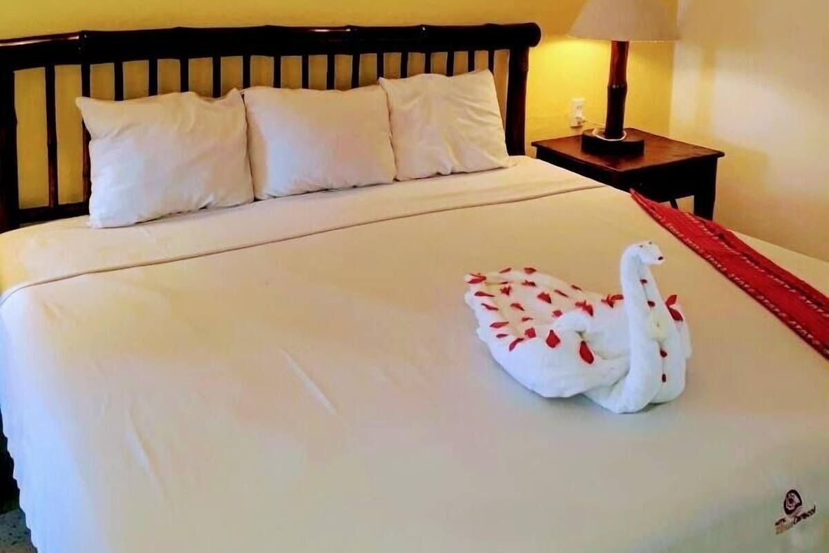 Habitación de hotel con cama, almohadas, mesita de noche con lámpara y un cisne decorativo sobre la cama.