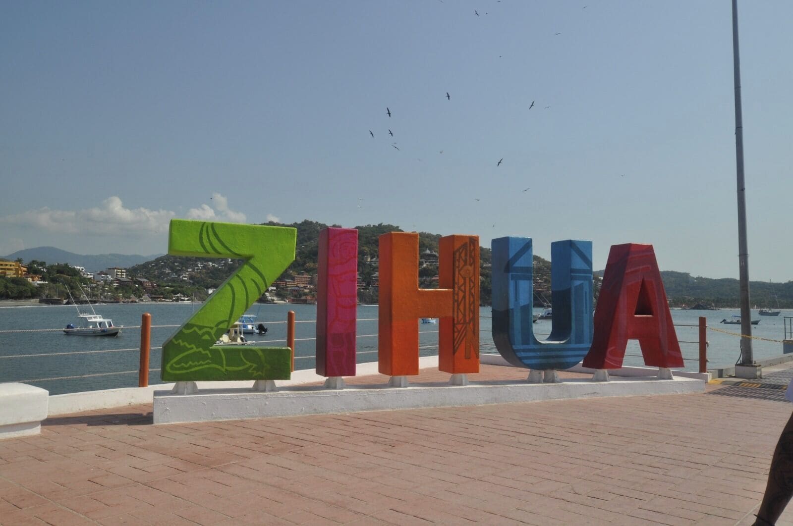 Un gran letrero al aire libre con la palabra 'ZIHUA' escrita con letras de colores, un sendero pavimentado y un cuerpo de agua con barcos al fondo.