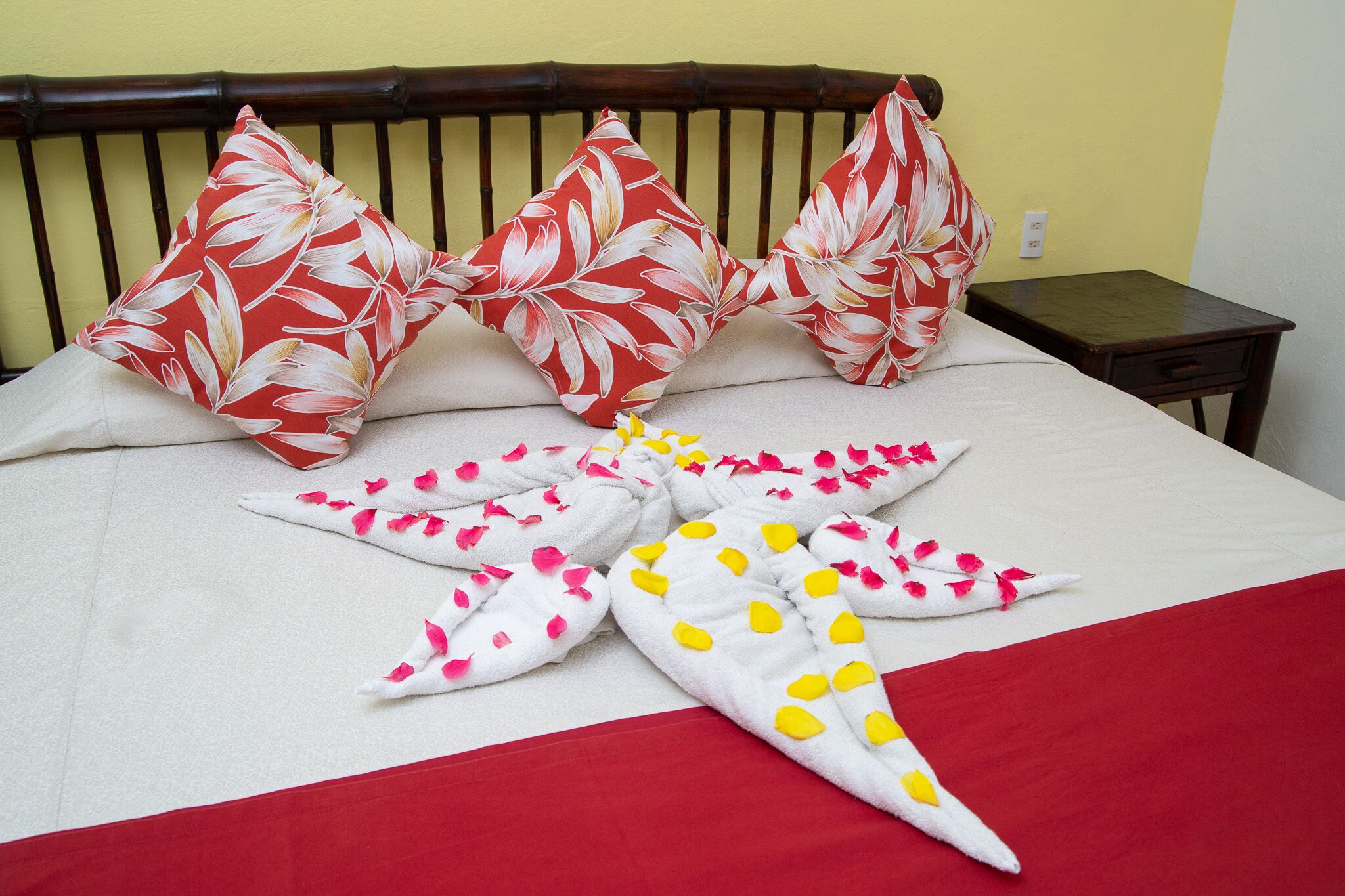 Una cama bien hecha con almohadas florales, una toalla blanca y una manta roja.