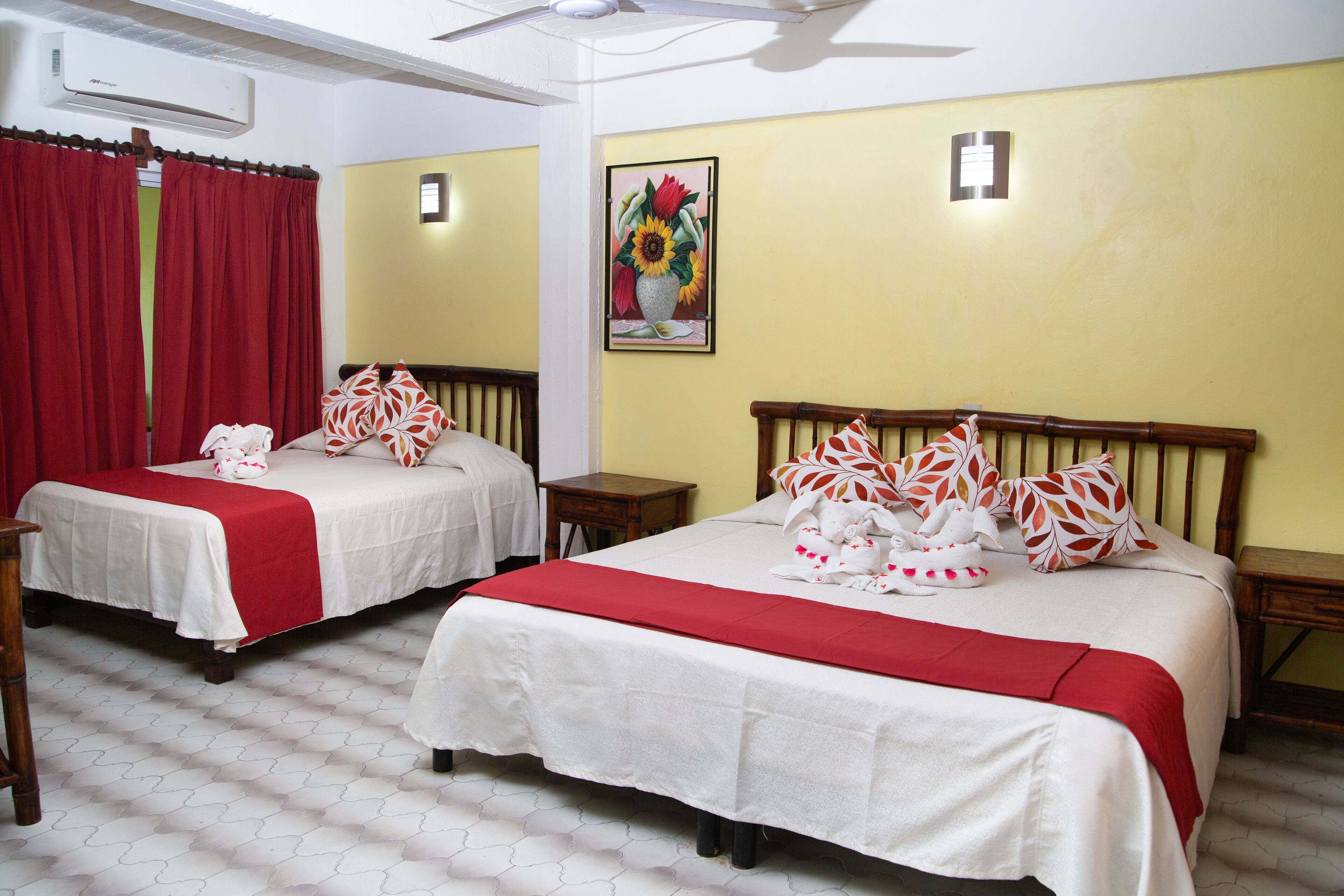 Dos camas individuales con ropa de cama roja y blanca, cabeceros de madera y un cuadro de girasoles en la pared amarilla.