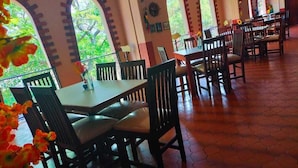 Un comedor con suelo de baldosas hexagonales, mesas y sillas de madera, y ventanas arqueadas con vistas a plantas y follaje.