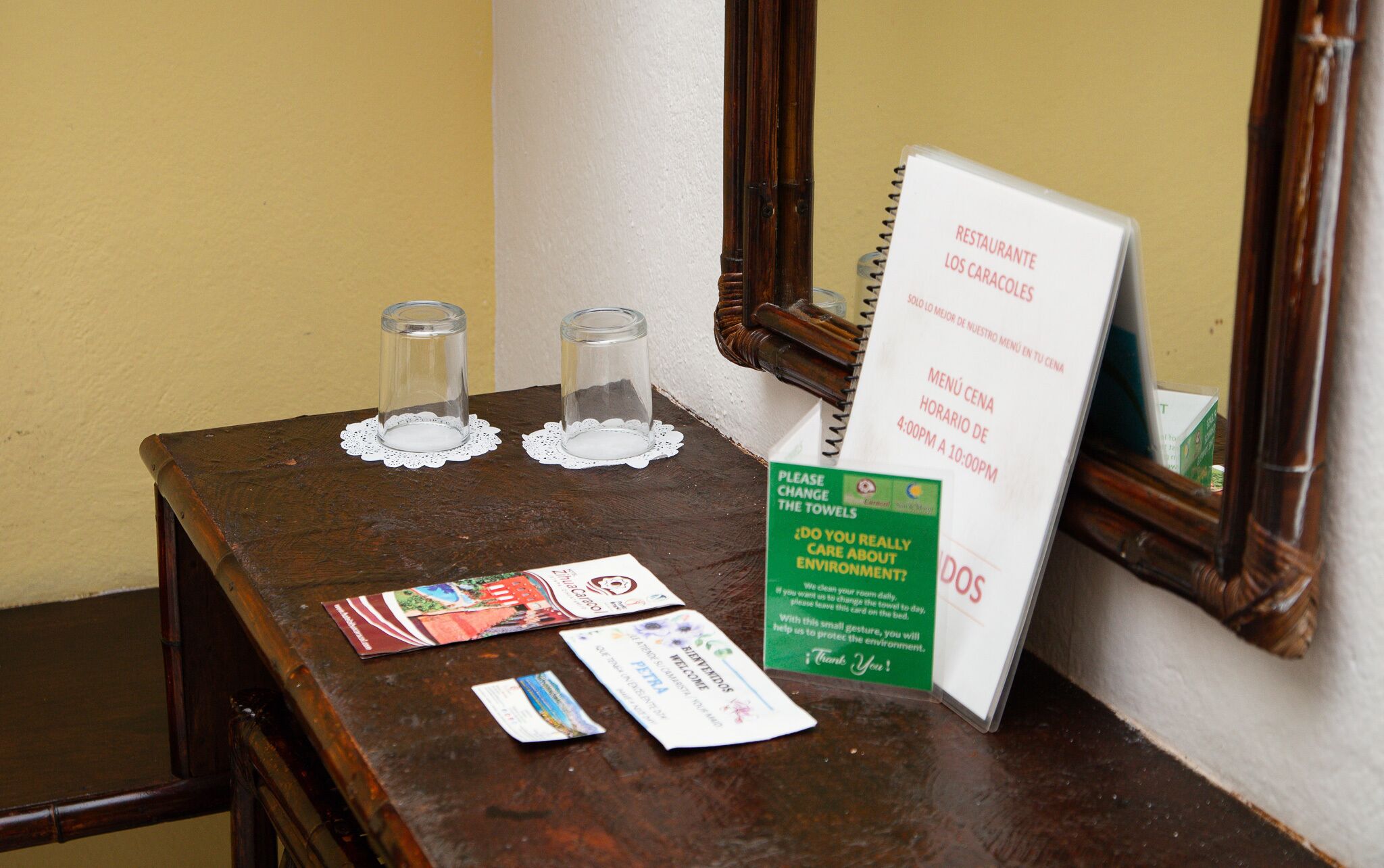 Una mesa de madera con dos vasos vacíos sobre servilletas de encaje, un letrero de menú y una tarjeta verde sobre cuidado del medio ambiente.