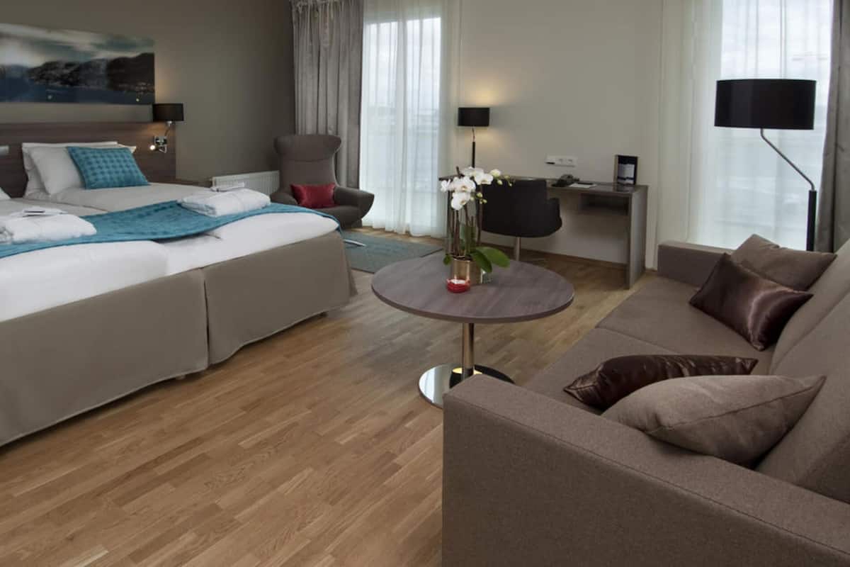 Habitación de hotel con cama, sofá, una mesita con un florero, un escritorio con lámpara y una silla.
