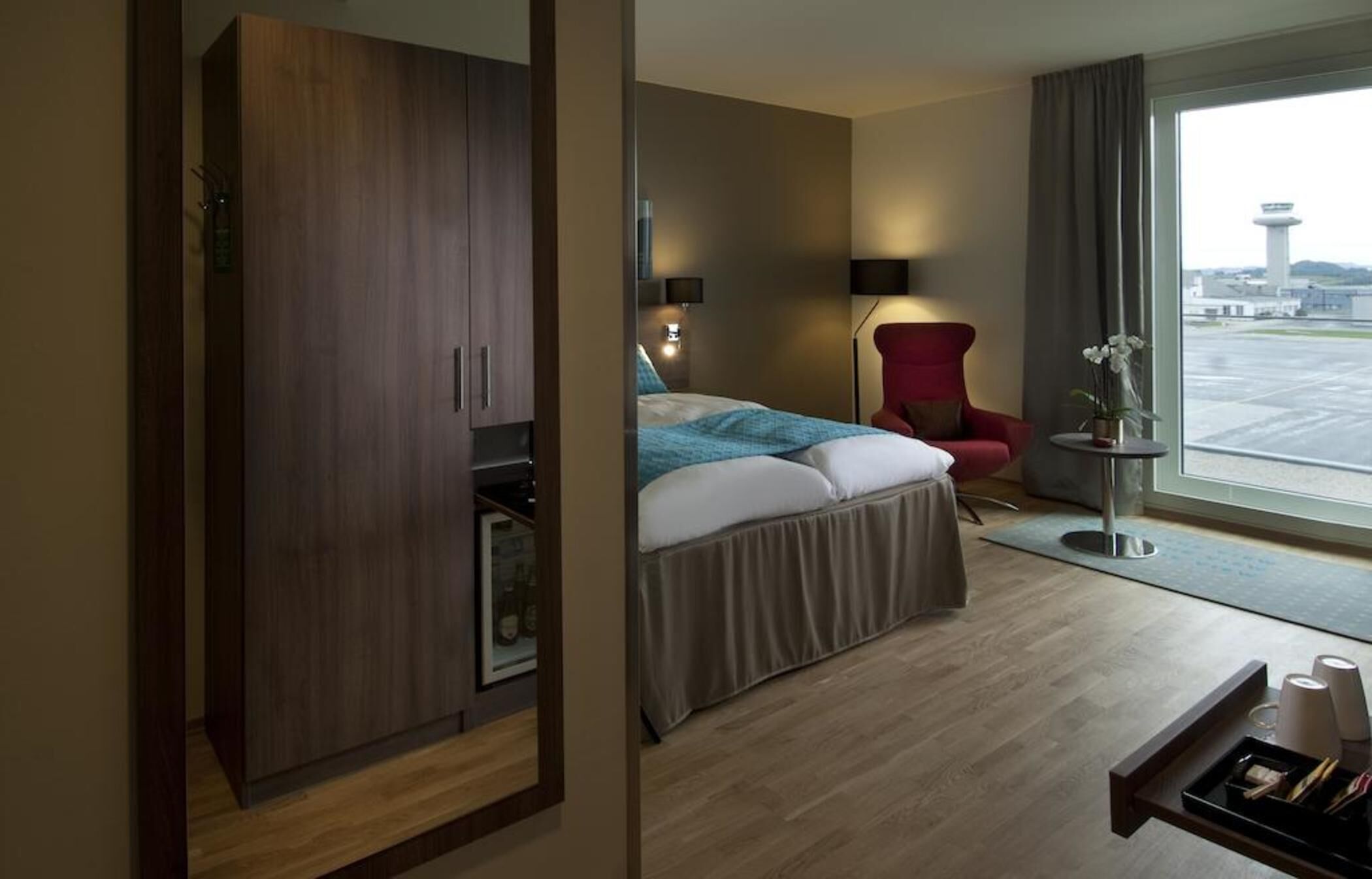 Et hotelværelse med en seng, en rød stol, et skrivebord med bakke og udsigt til en lufthavn.