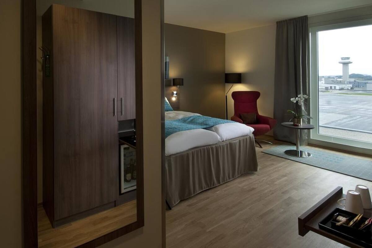 Habitación de hotel con cama, una silla roja, una mesita con bandeja y vistas al aeropuerto.