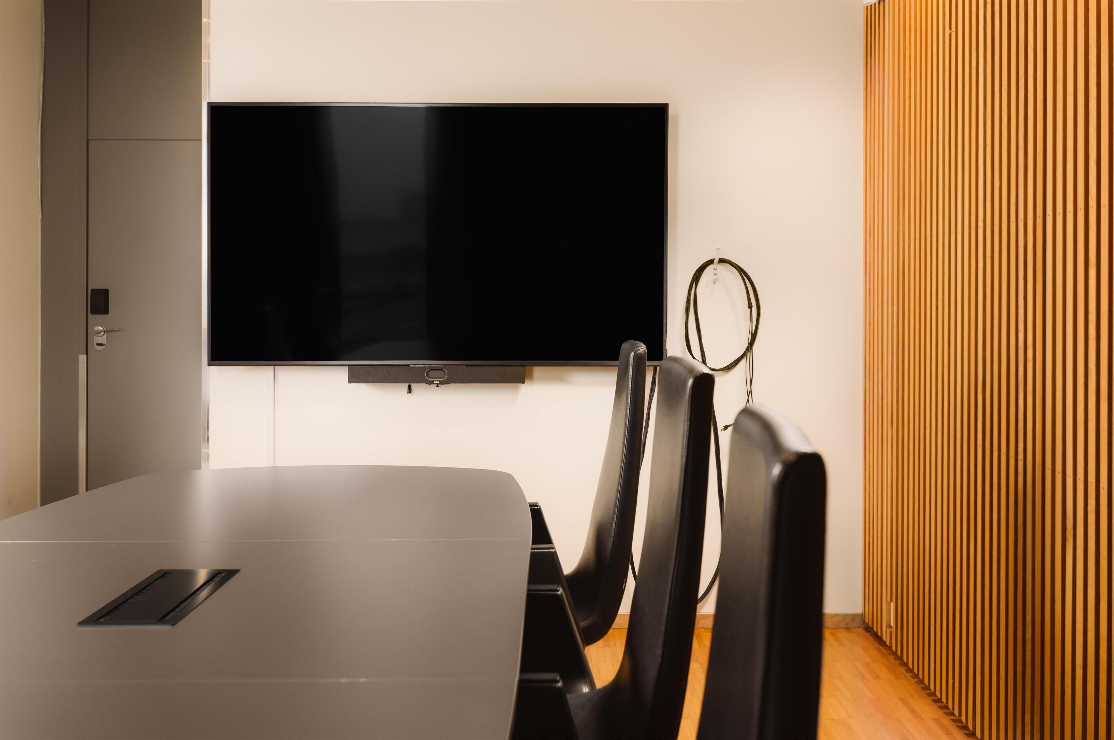 Een moderne vergaderruimte met een groot flatscreen aan de muur, een strakke tafel en zwarte stoelen.