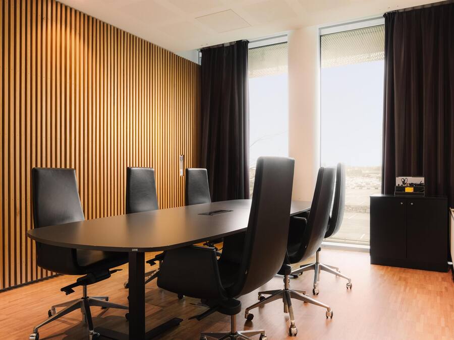 Ein Konferenzraum mit einem langen Tisch, sechs schwarzen Stühlen, einem schwarzen Schrank und einem Fenster mit Vorhängen.