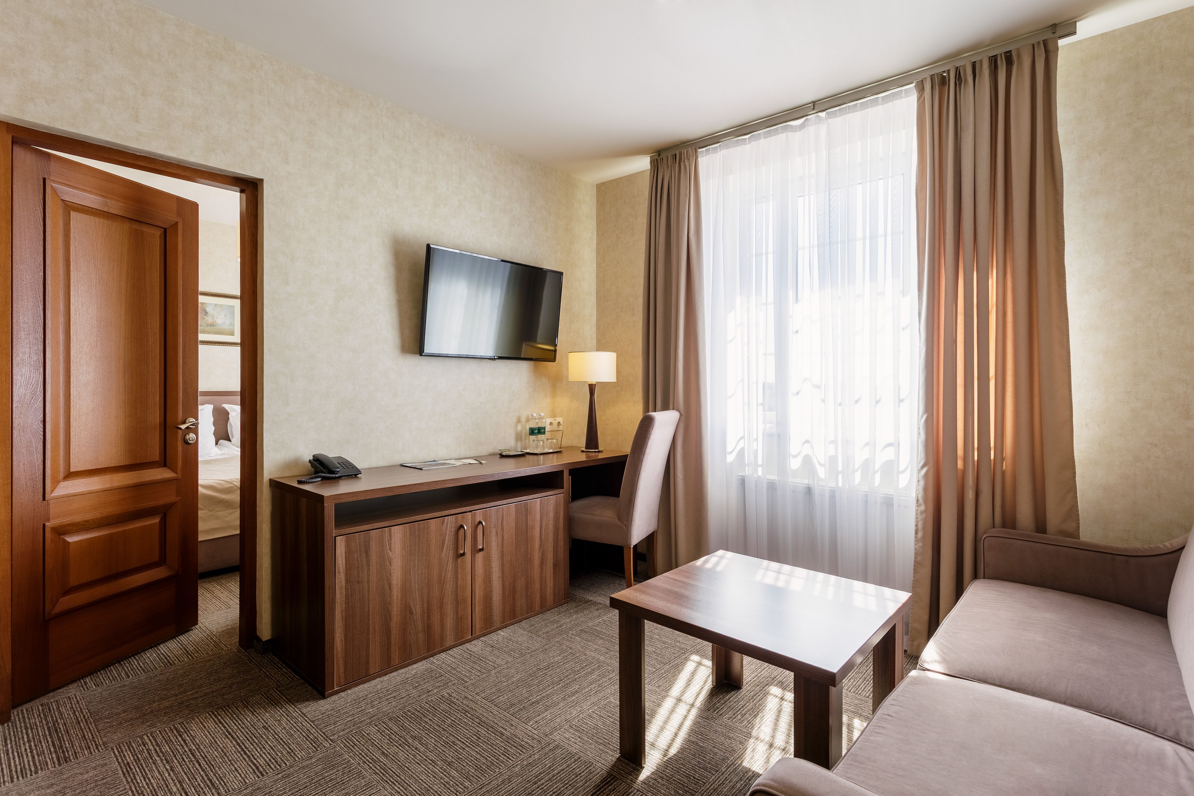 A hotel room with a bed, a desk with a telephone, a flat-screen TV, a lamp, a chair, a sofa, and a window with curtains.
