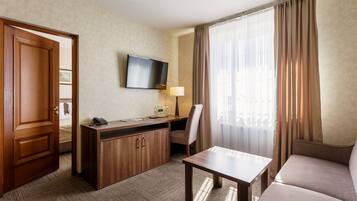 A hotel room with a bed, a desk with a telephone, a flat-screen TV, a lamp, a chair, a sofa, and a window with curtains.