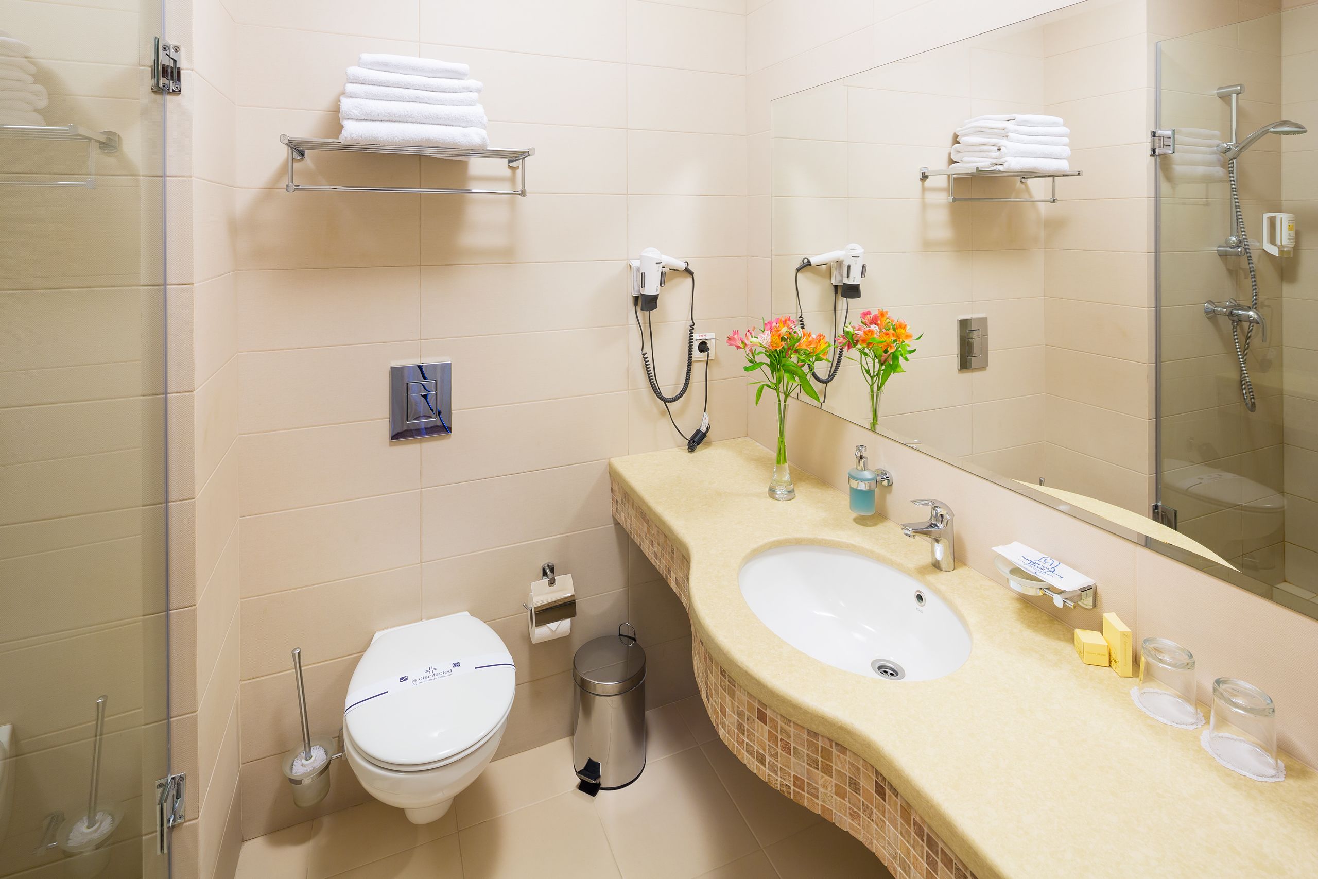 A bathroom with a toilet, sink, and shower. There are towels on shelves and a trash can.