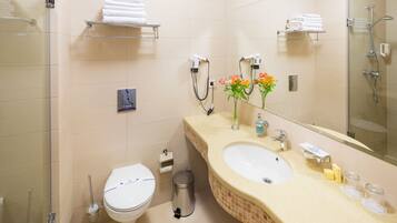 A bathroom with a toilet, sink, and shower. There are towels on shelves and a trash can.
