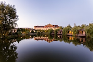 Un gran hotel con su reflejo en aguas tranquilas, rodeado de árboles y un pequeño puente.