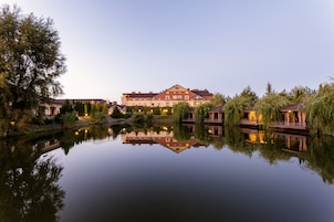 Un gran hotel con su reflejo en aguas tranquilas, rodeado de árboles y un pequeño puente.