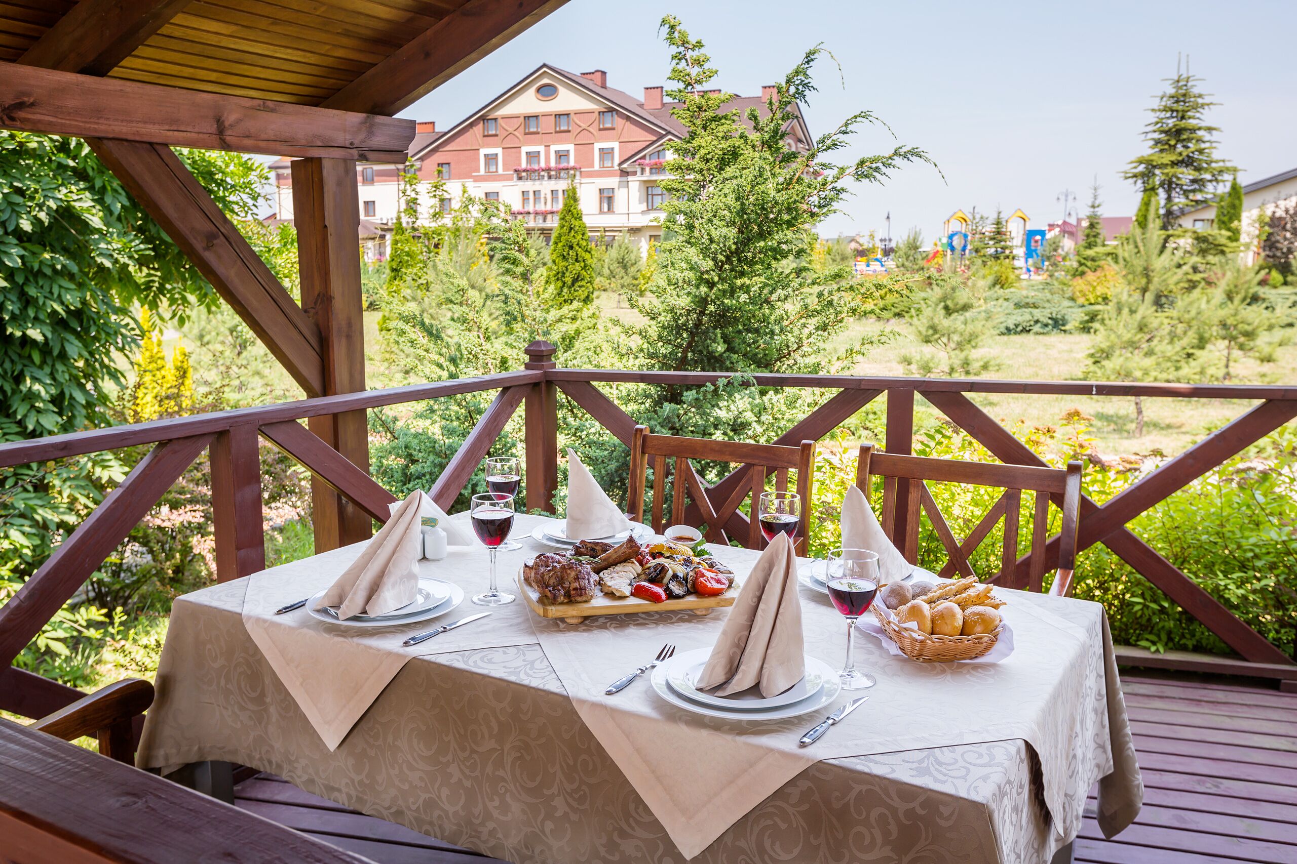 An outdoor dining area with a table set for a meal, surrounded by greenery and a view of a building in the background.