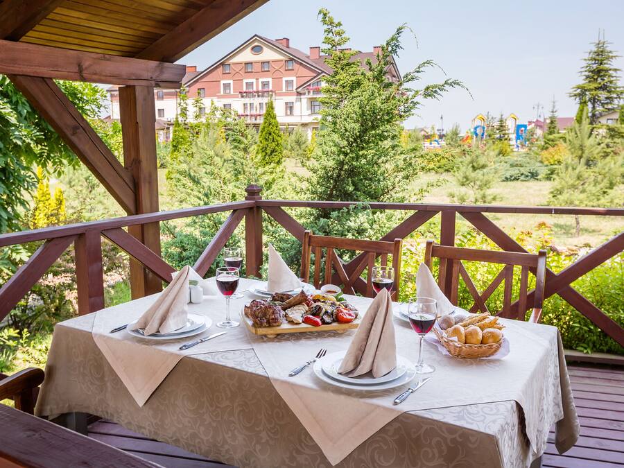 An outdoor dining area with a table set for a meal, surrounded by greenery and a view of a building in the background.