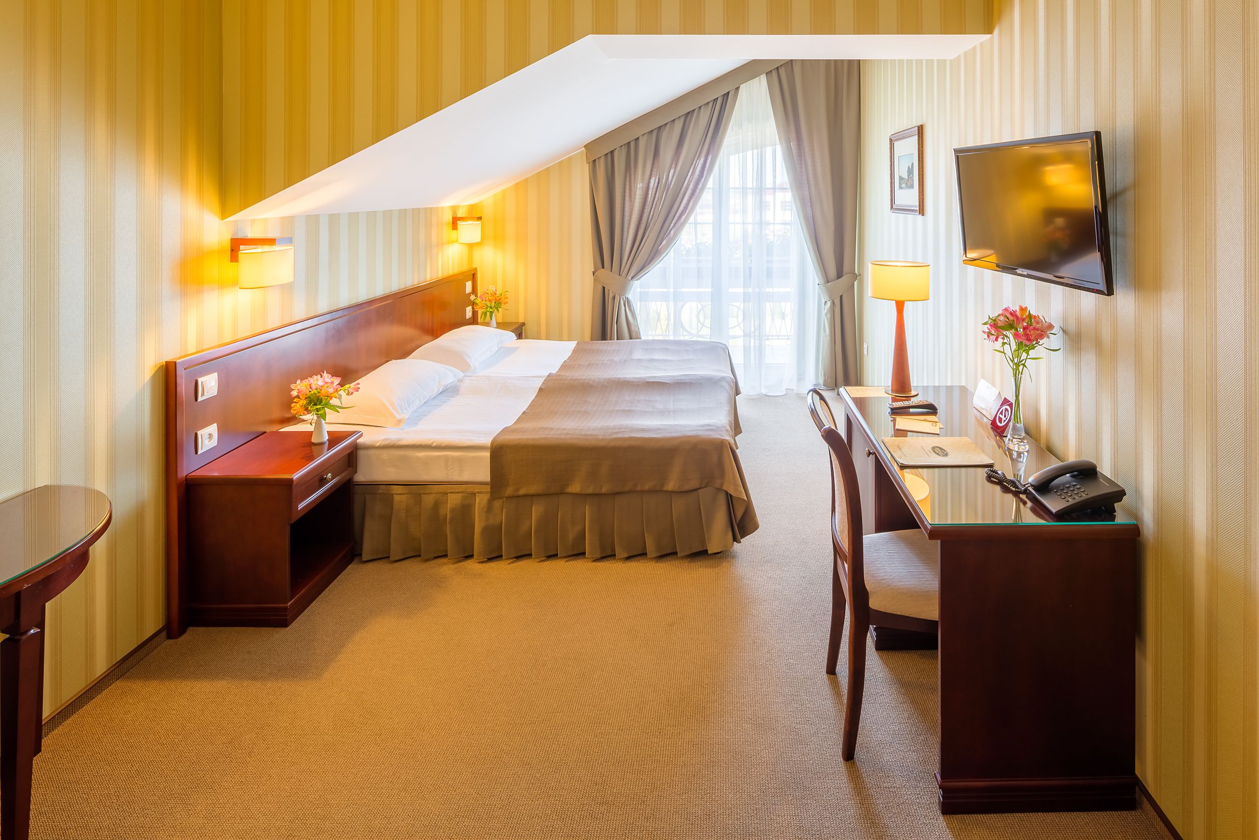 A hotel room with a large bed, bedside tables, a desk with a telephone, and a TV mounted on the wall.
