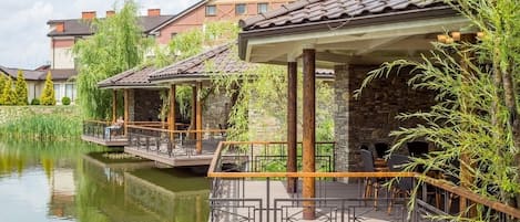 A riverside restaurant with outdoor seating, stone pillars, and a view of residential buildings.