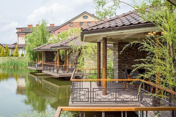 Un restaurante a orillas del río con terraza, pilares de piedra y vistas a edificios residenciales.