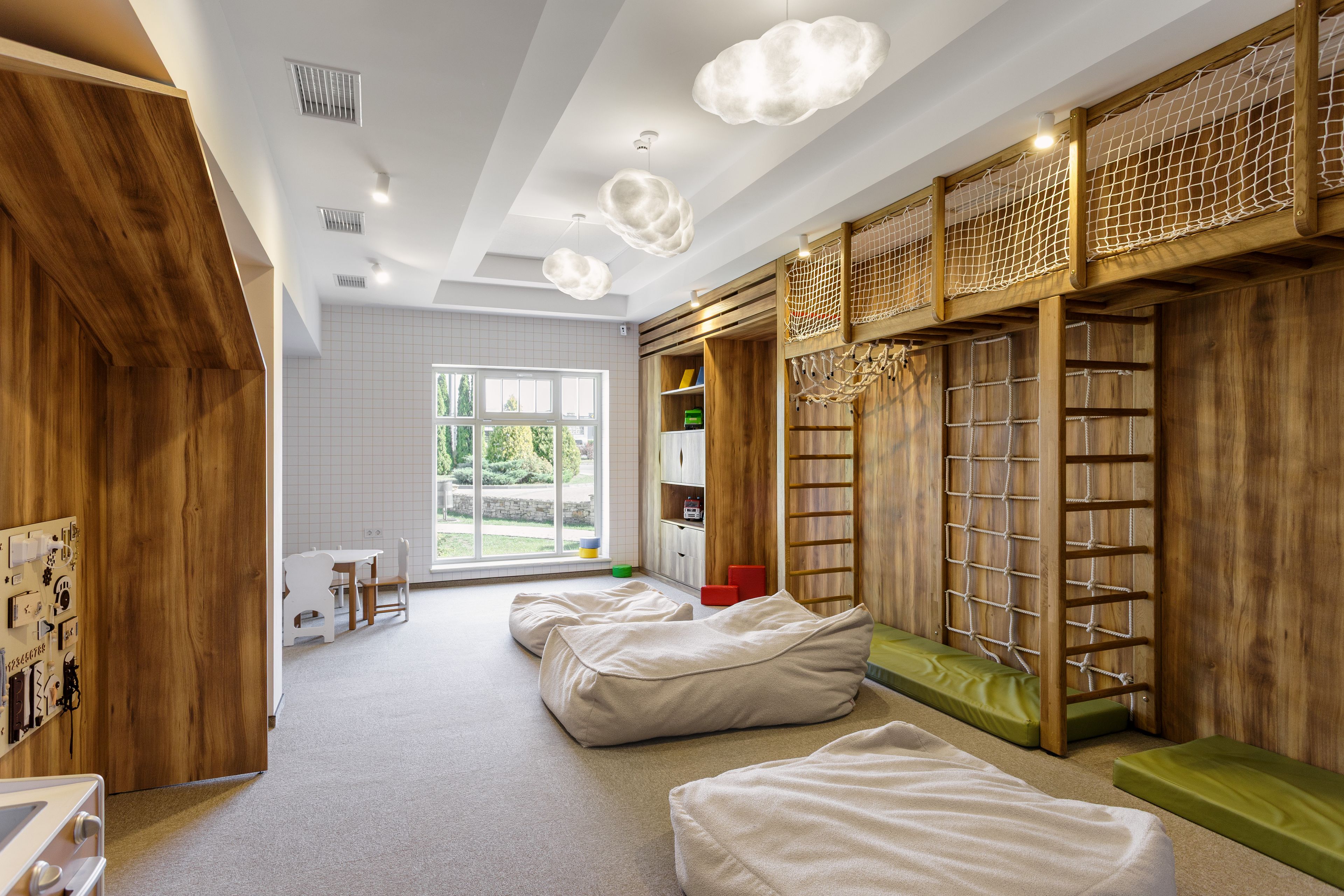 Um quarto com piso de madeira, uma janela, uma mesa branca com cadeiras e uma estante de livros.