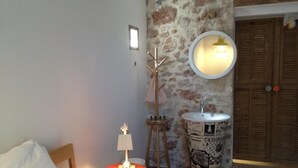 Un dormitorio con una cama, una mesa redonda con lámpara, un taburete de madera, un lavabo de diseño vintage y una pared de piedra.