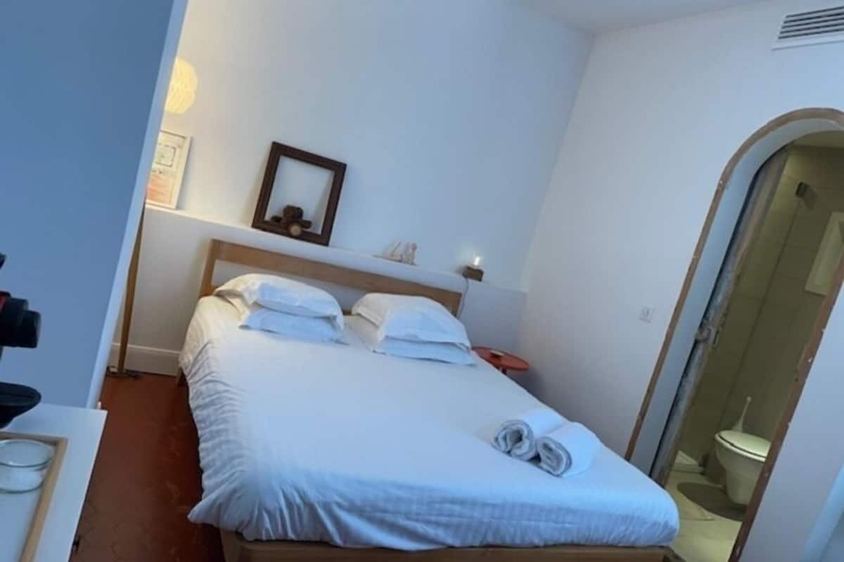 Ein ordentlich gemachtes Bett mit weißen Leinen und einem auf dem Bett gefalteten Handtuch, ein Nachttisch mit einer Lampe und ein Spiegel an der Wand.