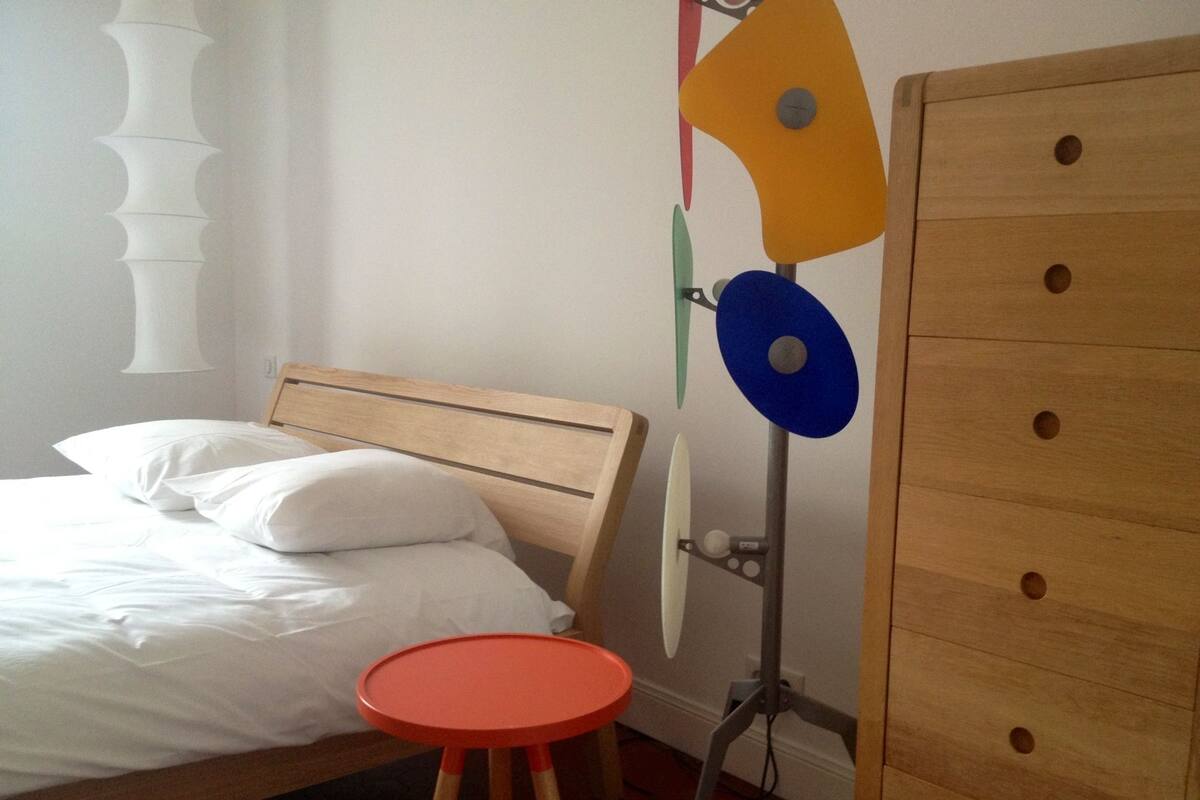 Ein Schlafzimmer mit einem hölzernen Bett, weißen Kissen, einem roten Hocker und einer Kommode aus Holz mit mehreren Schubladen.