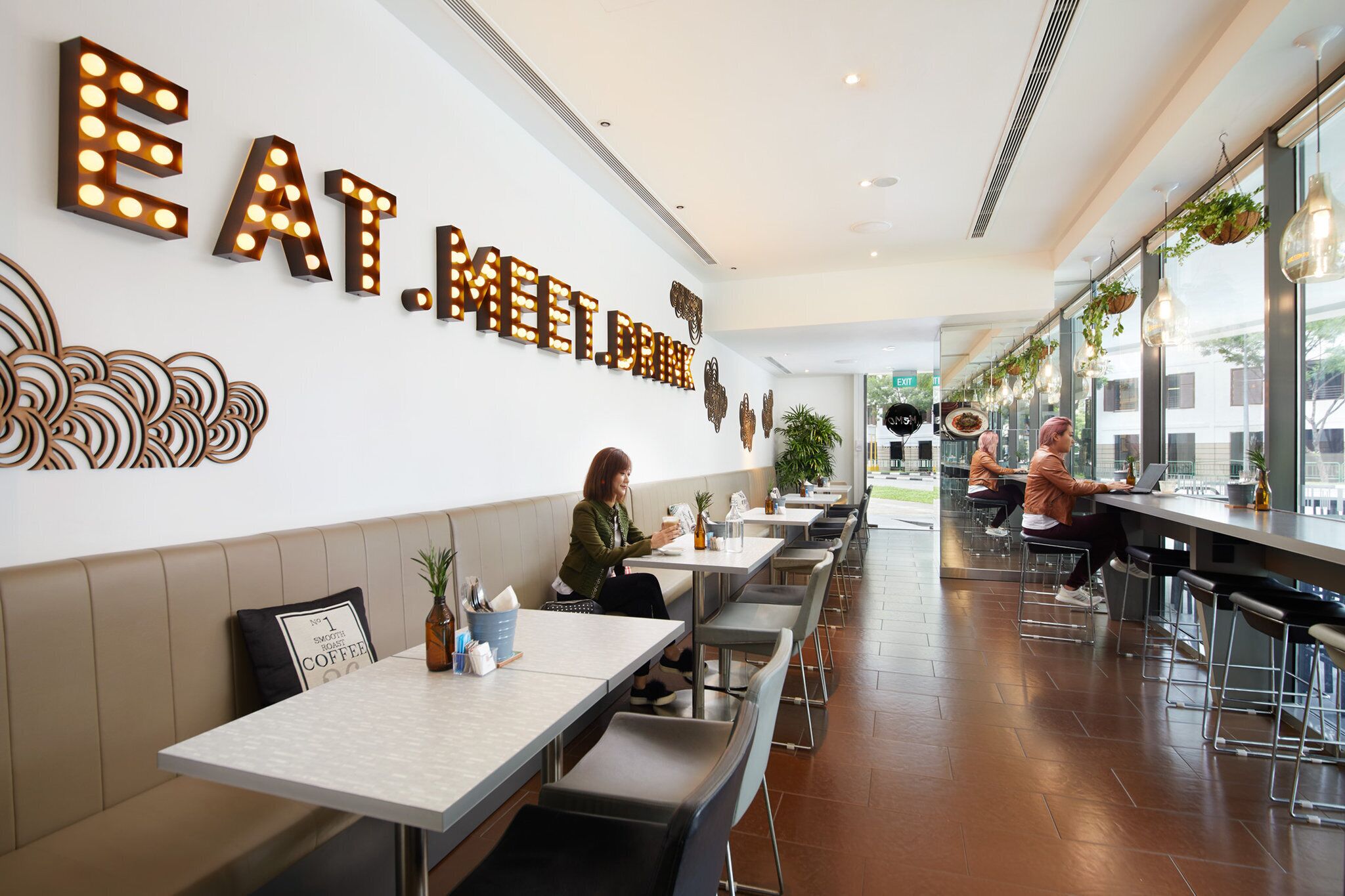 Un café moderno con una barra larga y mesas con asientos, con un cartel iluminado que dice 'EAT. MEET. DRINK.' y una pieza de arte decorativo en la pared.