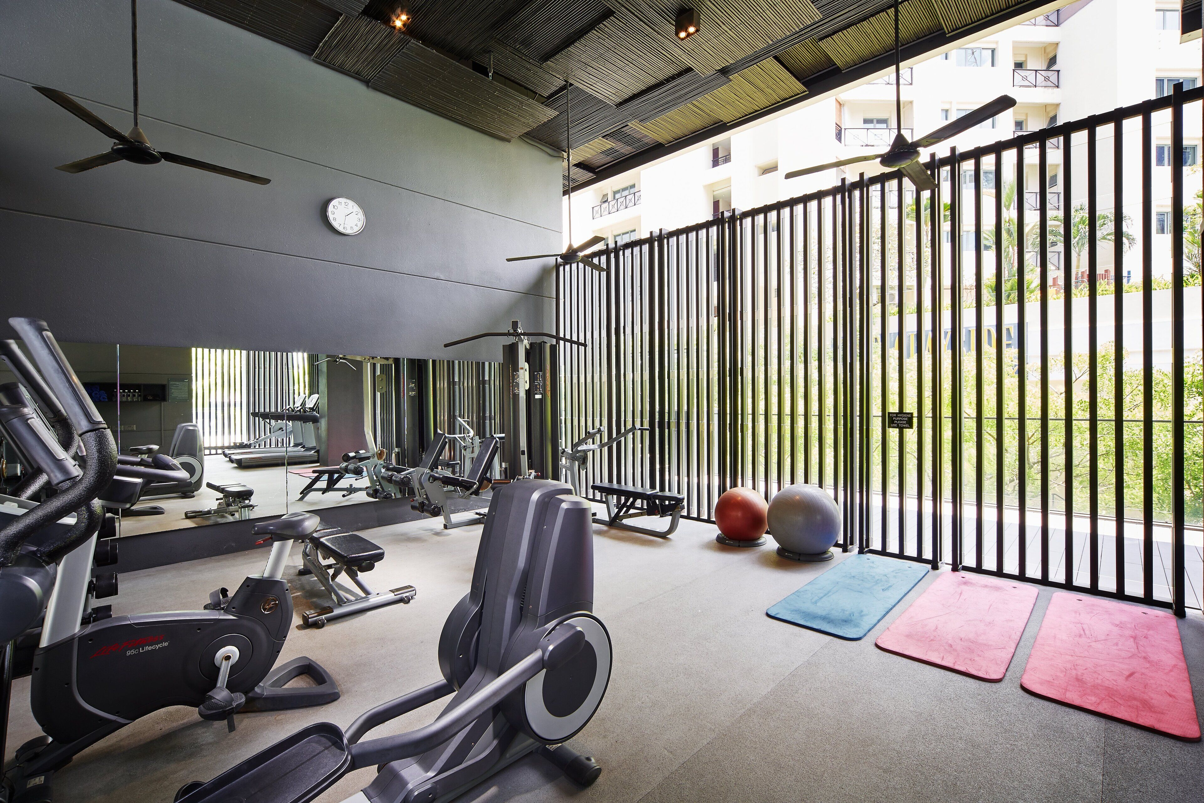 A gym with exercise equipment, a clock, and a view of a building through a fence.