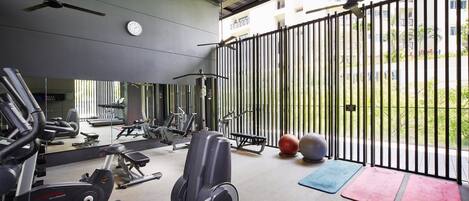 A gym with exercise equipment, a clock, and a view of a building through a fence.