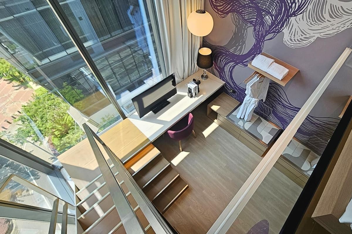 A modern room with a desk, a chair, a lamp, and a large window overlooking a cityscape.