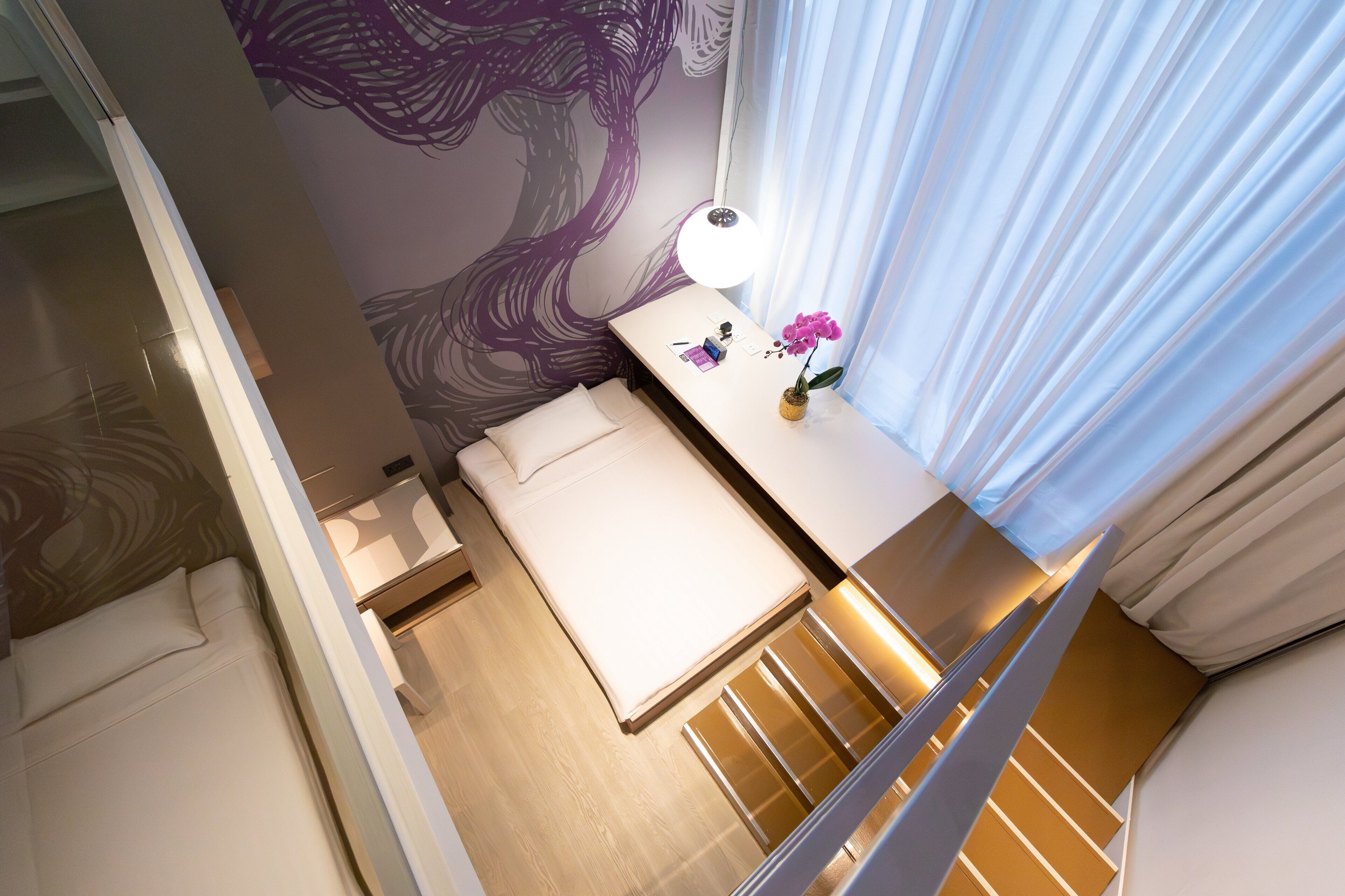 A modern hotel room with a bed, a desk, a lamp, and a window with curtains.