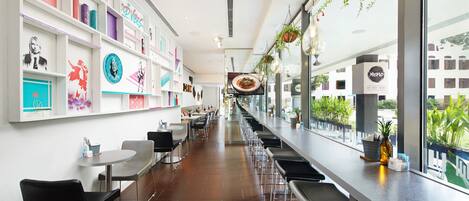 Un restaurante moderno con una barra larga, sillas negras y una pared decorada con obras de arte coloridas.