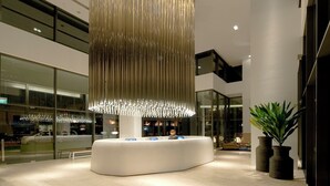 A modern lobby with a central reception desk, a large hanging light fixture, and a view of the outside through large windows. - Studio M Hotel Singapore (Singapore)