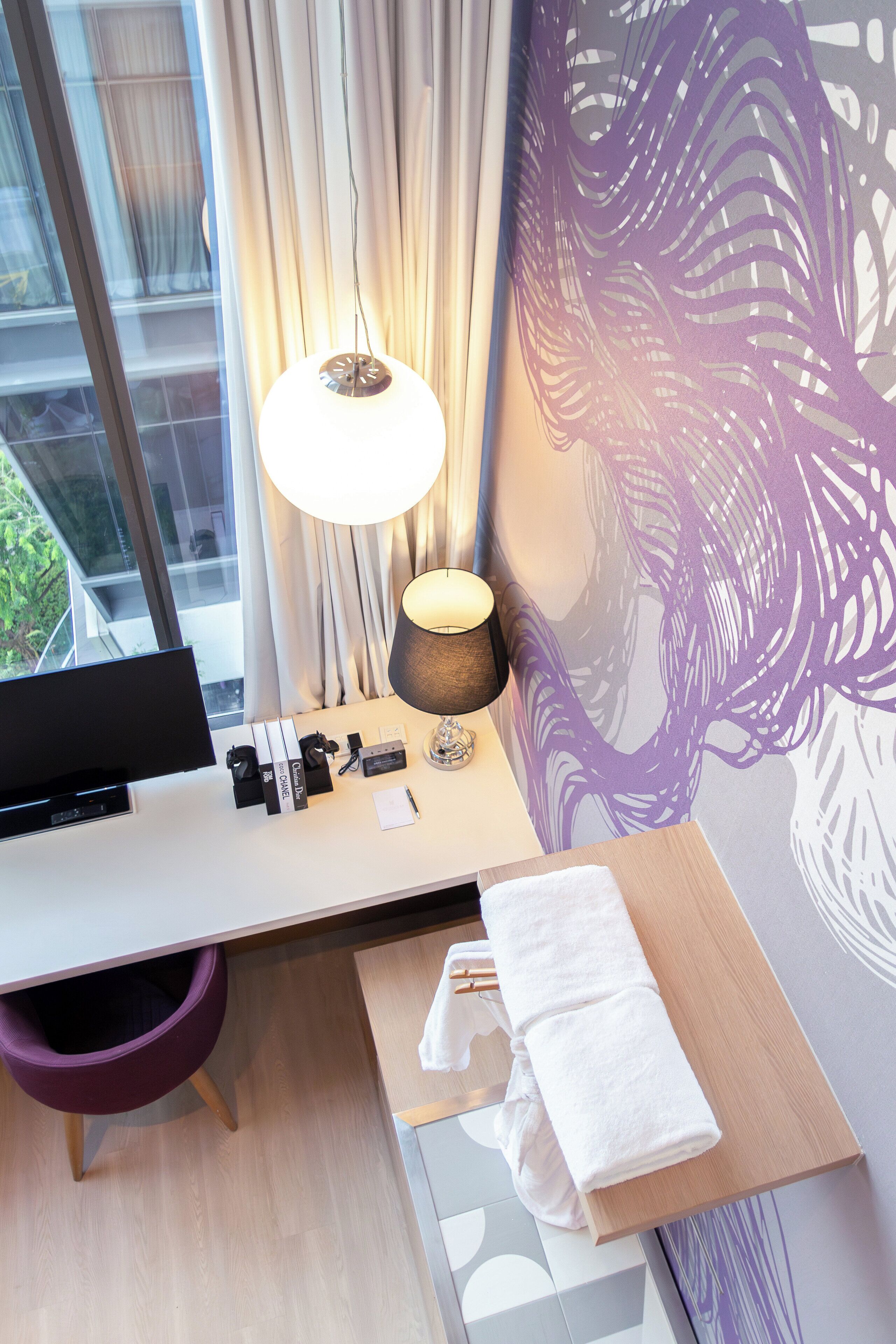 A hotel room with a desk, a lamp, a TV, a window with curtains, and a wall with a purple abstract design.