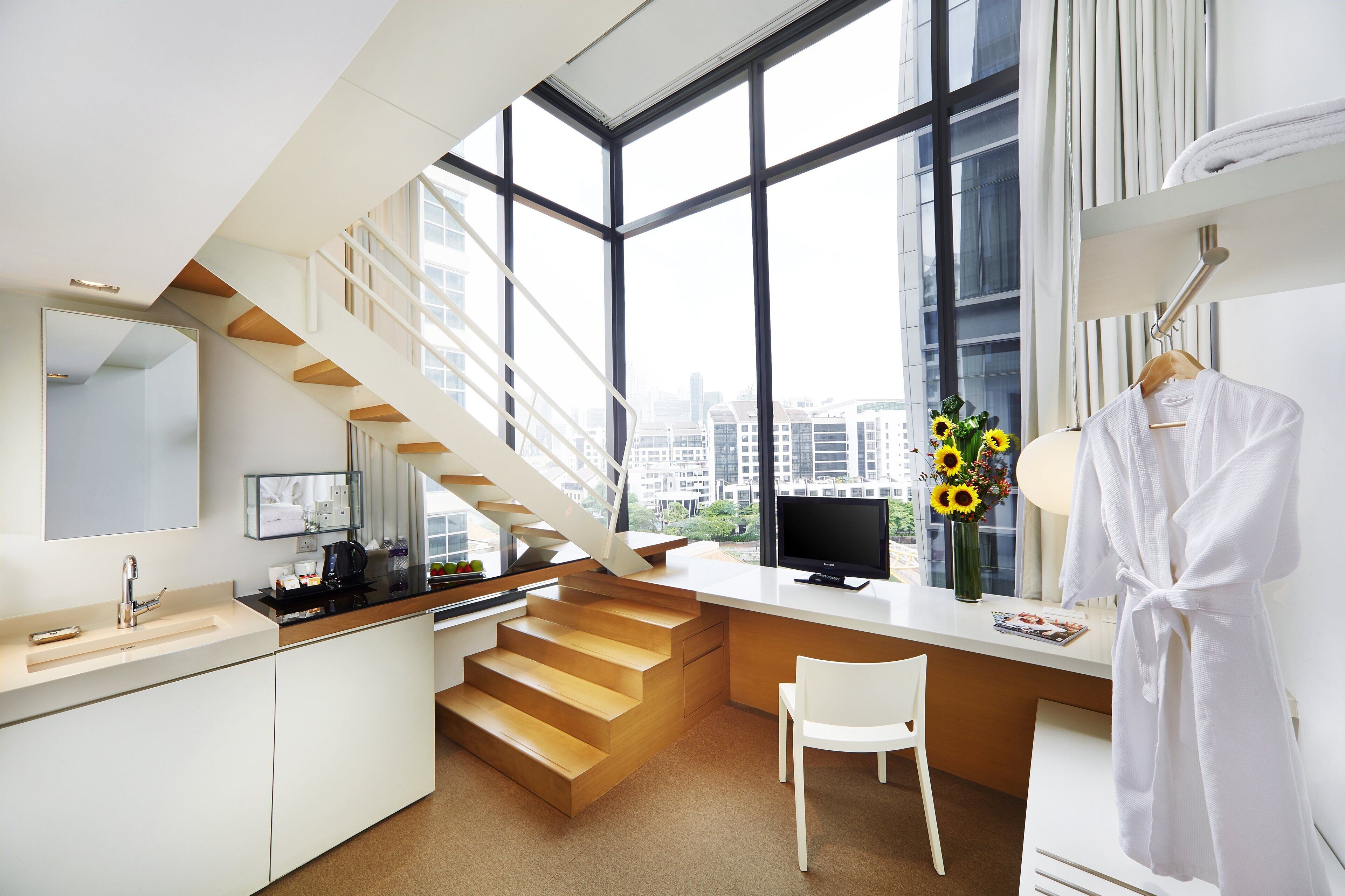 A modern hotel room with a kitchen area, a white chair, a desk, a large window with a city view, and a robe hanging on the wall.