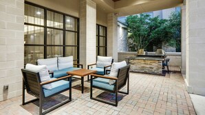 An outdoor patio with a brick floor, two sofas with cushions, a wooden table, and a stone fireplace. - Homewood Suites Dallas Allen (Allen)