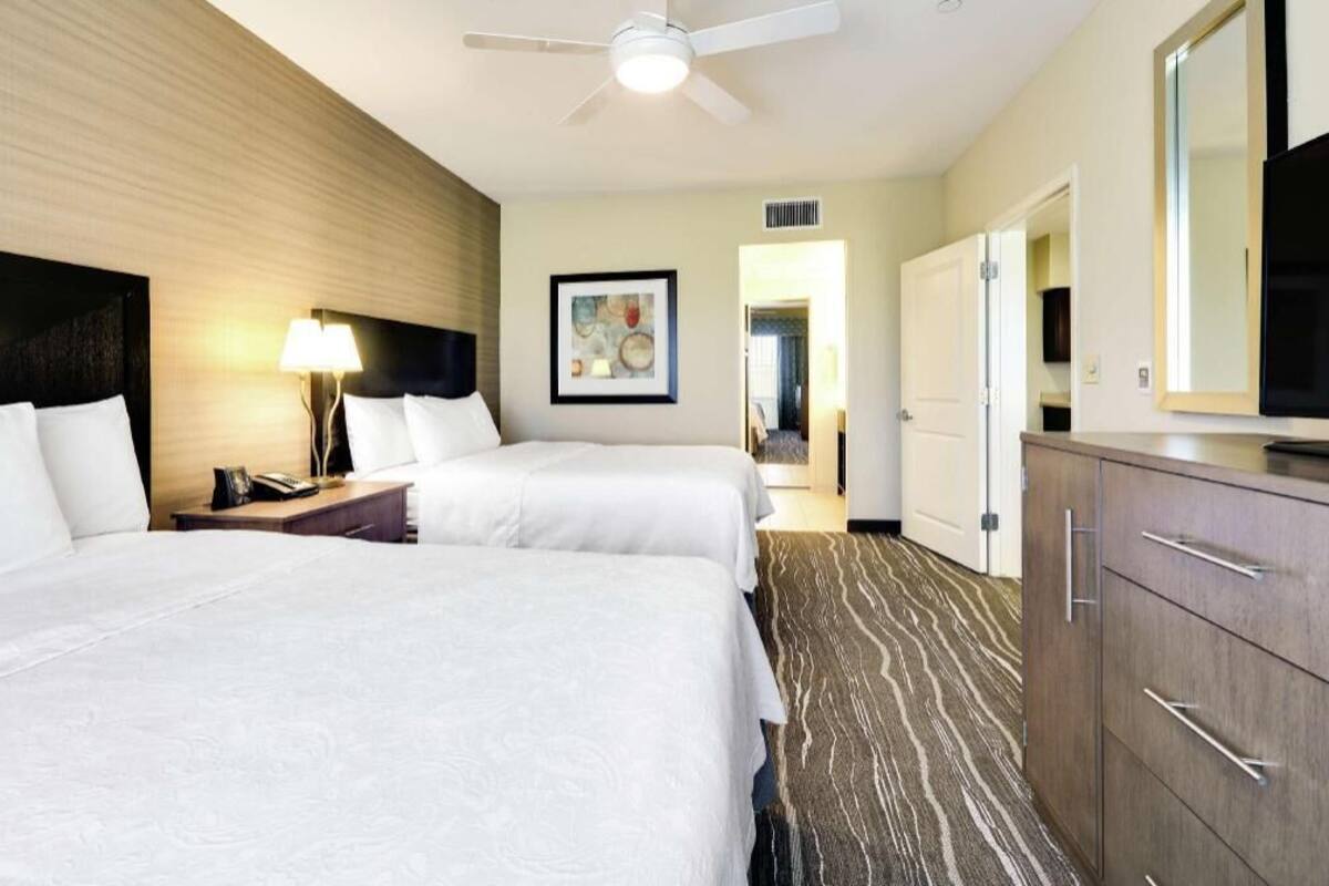 Une chambre d’hôtel avec deux lits, un ventilateur de plafond, un bureau avec une lampe, une télévision et une armoire.