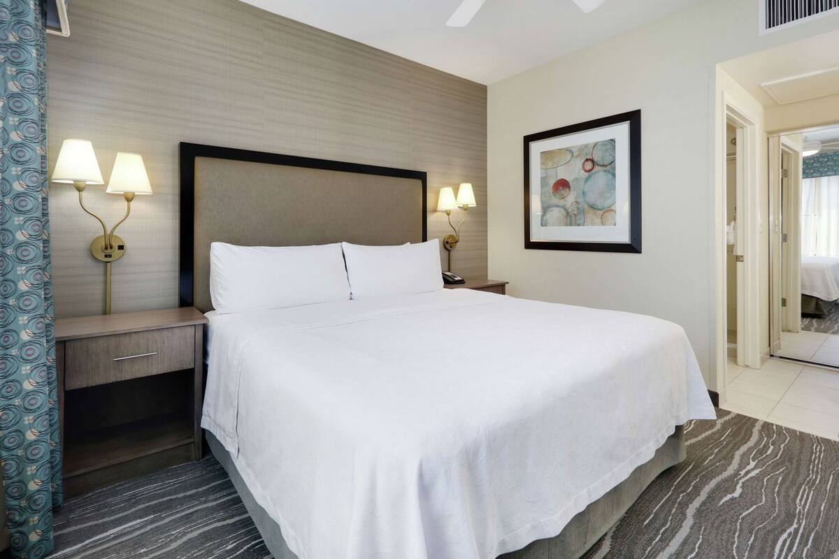 Une chambre d’hôtel comprenant un lit, des tables de chevet, des lampes, un tableau et une porte donnant sur une salle de bain.