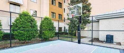 An outdoor basketball court with a chain-link fence, trees, and a building in the background.