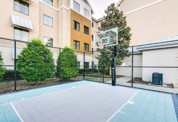 An outdoor basketball court with a chain-link fence, trees, and a building in the background.