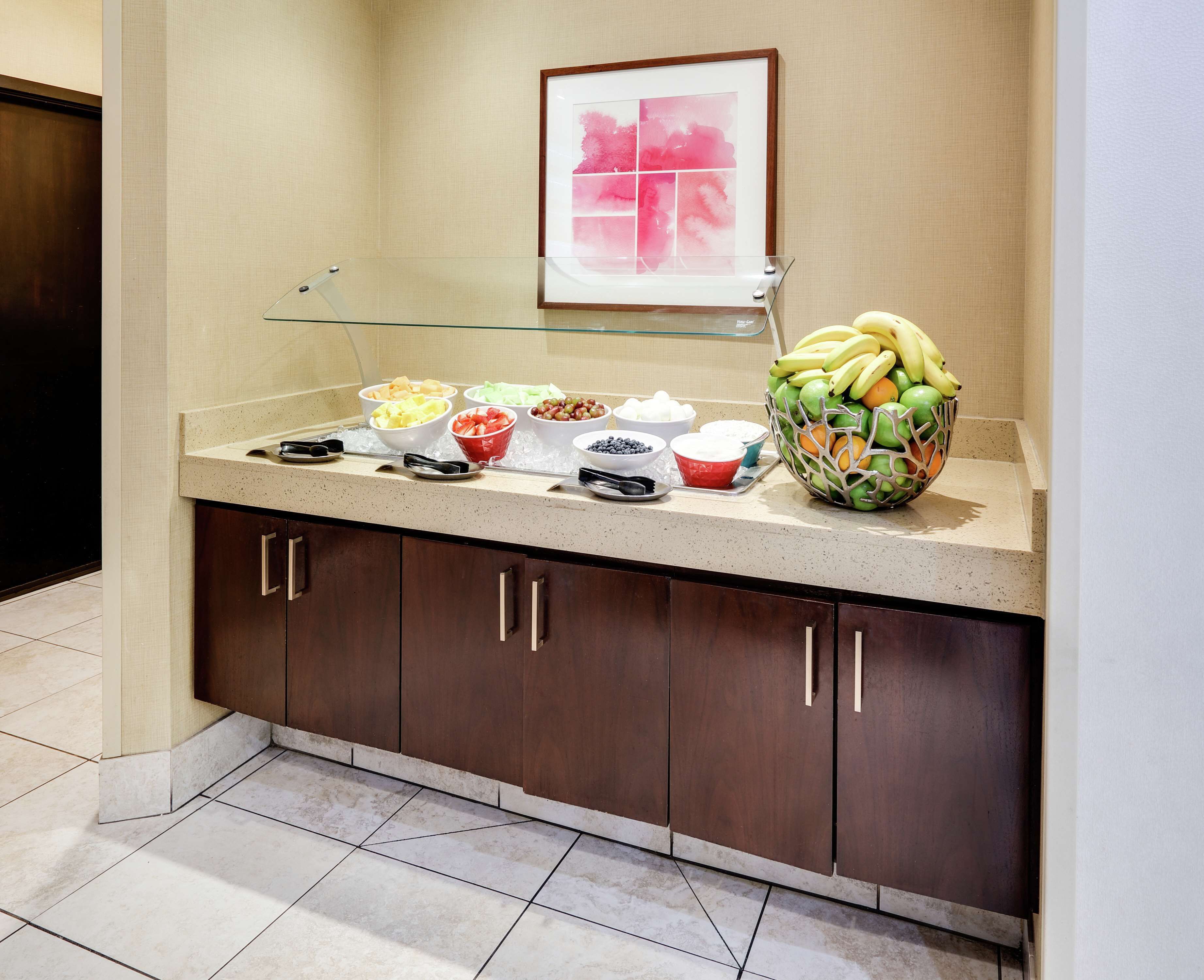 A buffet counter with various fruits and bowls of food, a framed picture on the wall, and a tiled floor.