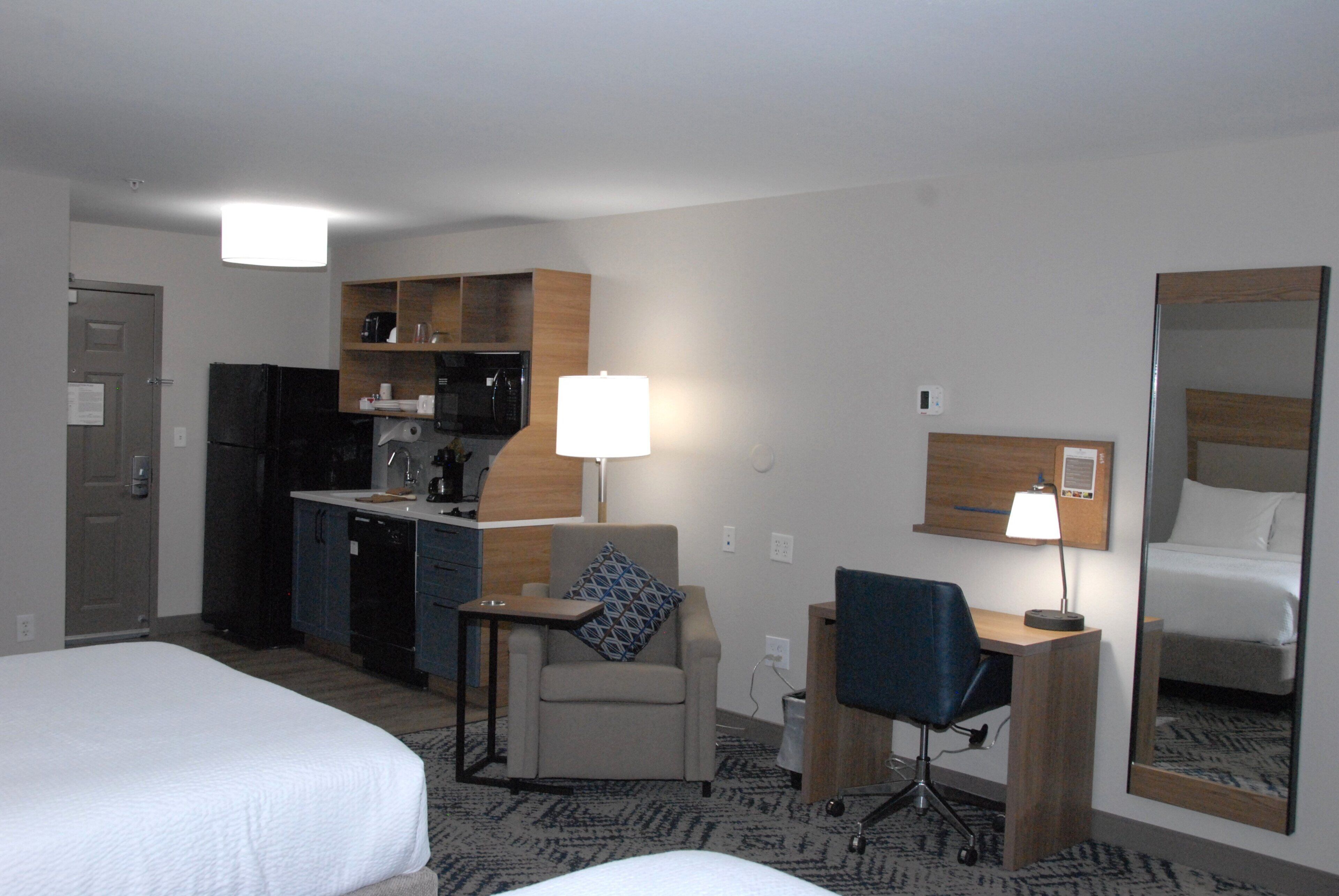 A hotel room with a bed, a desk, a chair, a kitchenette, and a mirror.